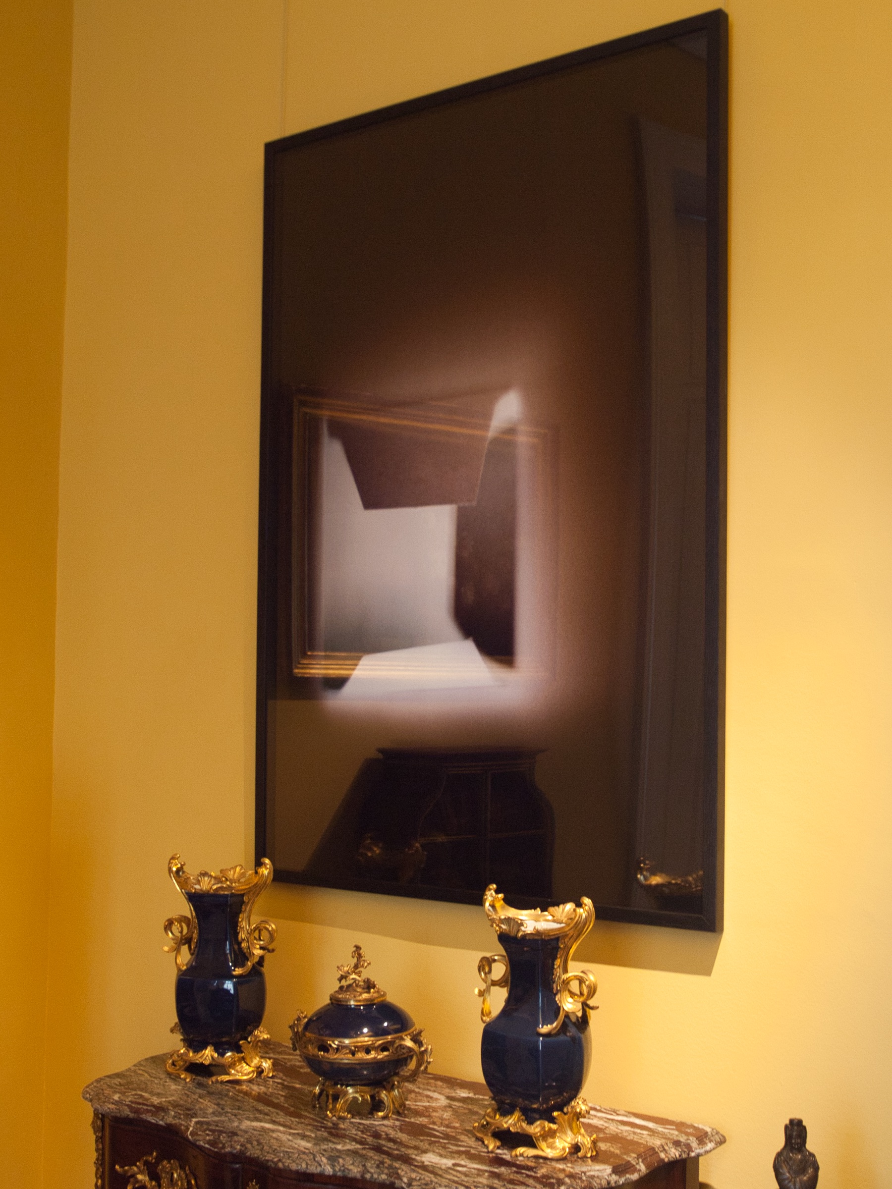 L'image montre un intérieur élégant avec des murs peints en jaune. Il y a un grand tableau encadré en noir accroché au mur, représentant une composition abstraite de lumière et d'ombre. En dessous du tableau, on peut voir une console ornée en bois avec un marbre. Sur cette console, deux vases en céramique bleue avec des embellissements dorés sont positionnés de manière symétrique, ainsi qu'un autre objet décoratif en forme de boîte, également en bleu et or. L'ensemble crée une atmosphère sophistiquée et artistique.