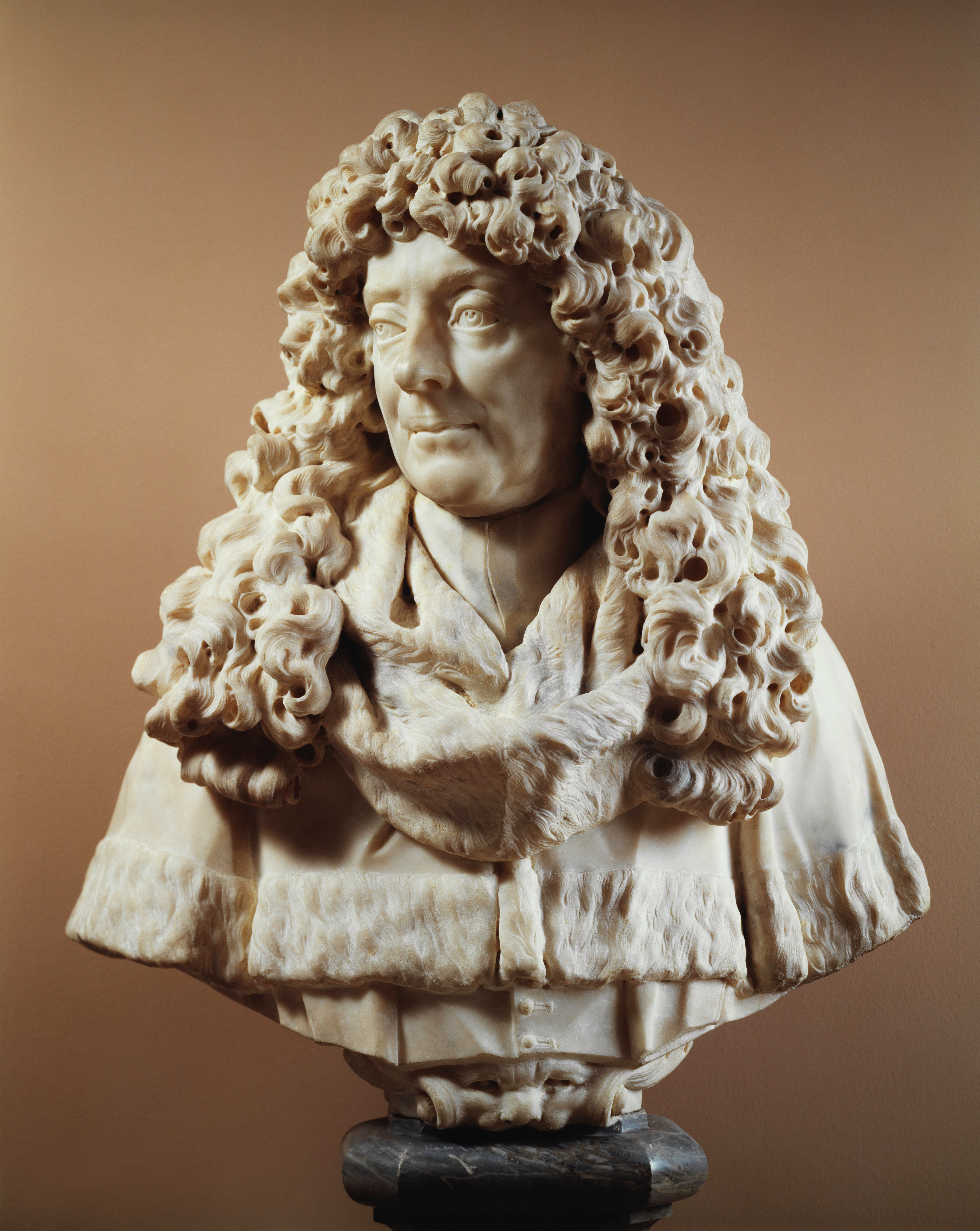 Cette image représente un buste sculpté en marbre d'un homme au visage expressif. Il a de longs cheveux bouclés, ornés de mèches détaillées, et porte un manteau ou une écharpe qui lui donne une allure élégante. Le buste est bien travaillé, montrant une attention particulière aux détails du visage et des cheveux, et il est placé sur un socle noir qui contraste avec la couleur claire du marbre. L'arrière-plan est de couleur neutre, ce qui met en valeur la sculpture.
