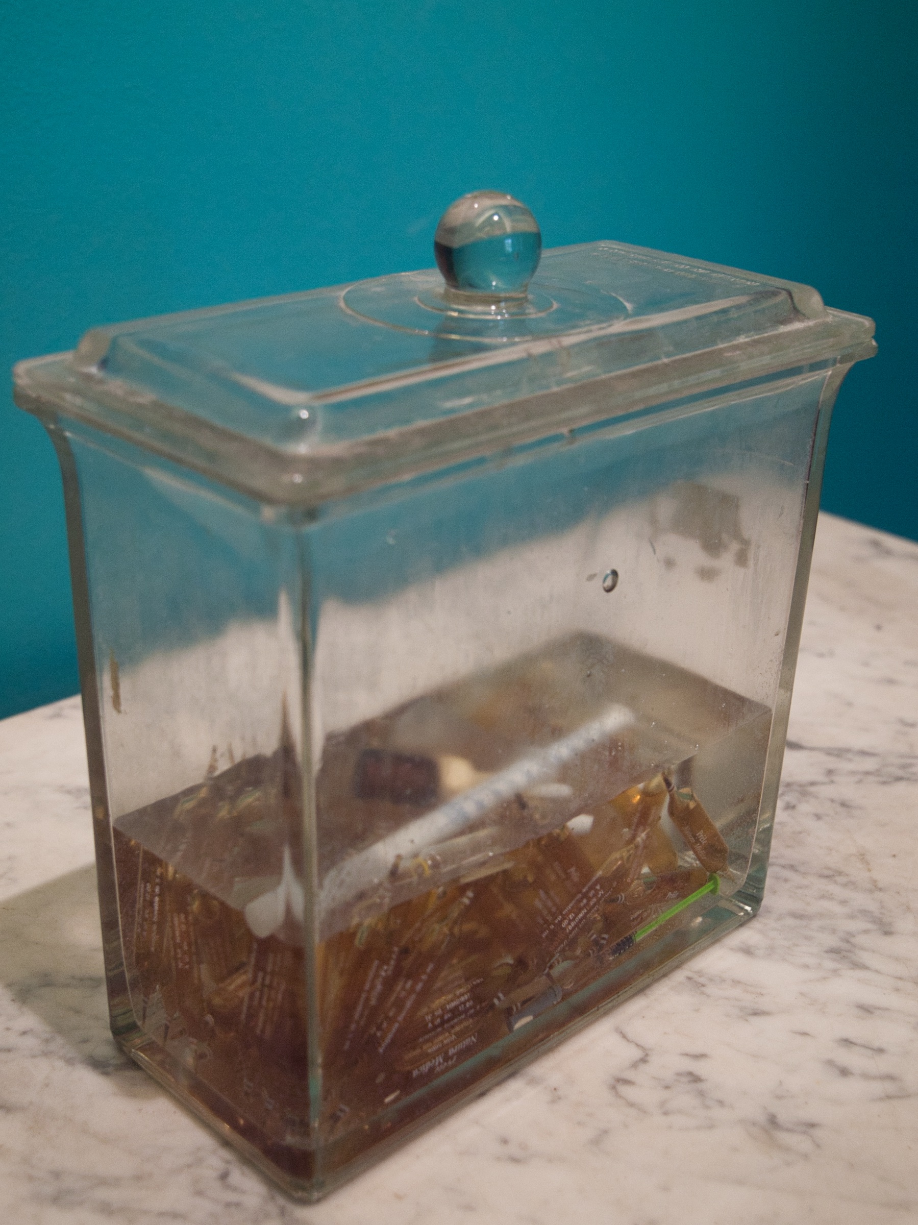 L'image montre un bocal en verre transparent avec un couvercle, qui est rempli de petits morceaux, probablement des bonbons ou des sucreries. Le bocal est placé sur une surface en marbre, et en arrière-plan, on peut voir un mur de couleur turquoise. La lumière semble éclairer le bocal, mettant en évidence la transparence du verre et le contenu à l'intérieur.