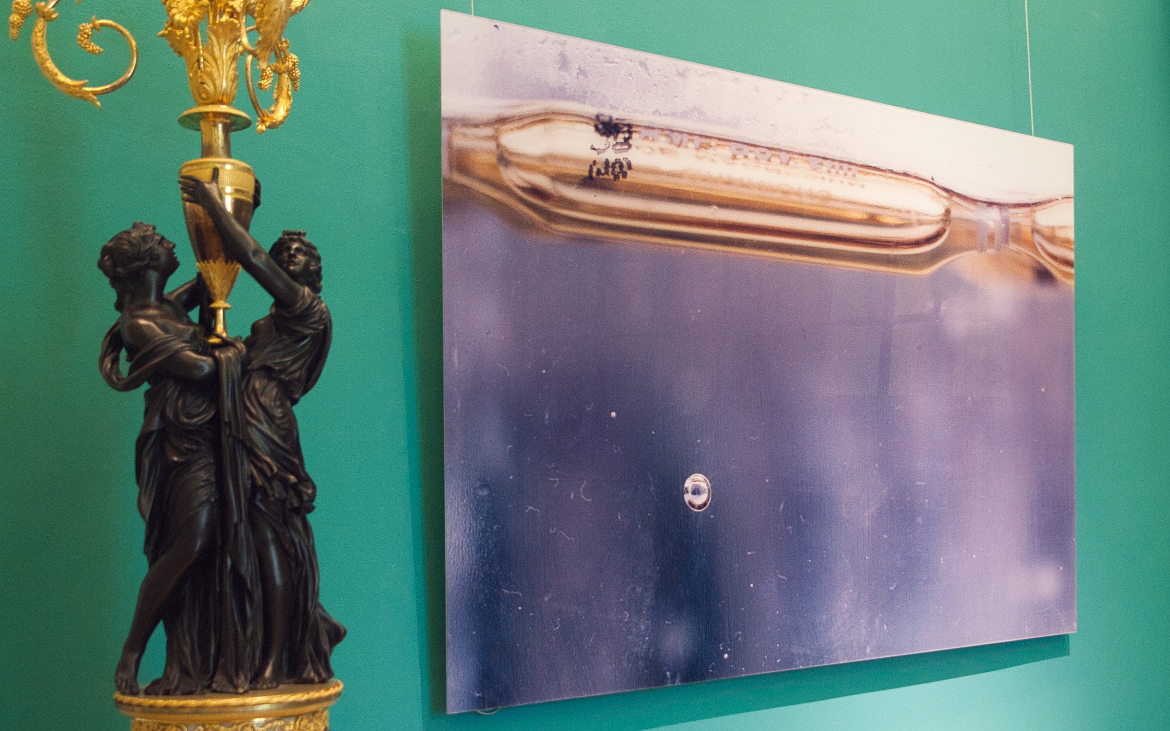 L'image présente une œuvre d'art accrochée au mur d'une pièce aux murs verts. L'œuvre semble être une photographie abstraite, probablement d'une surface réfléchissante ou d'un liquide, avec des nuances de bleu et des détails subtils. À côté, il y a une sculpture en or, représentant deux figures humaines qui se tiennent l'une l'autre. Cela crée un contraste intéressant entre l'art contemporain et la sculpture classique.