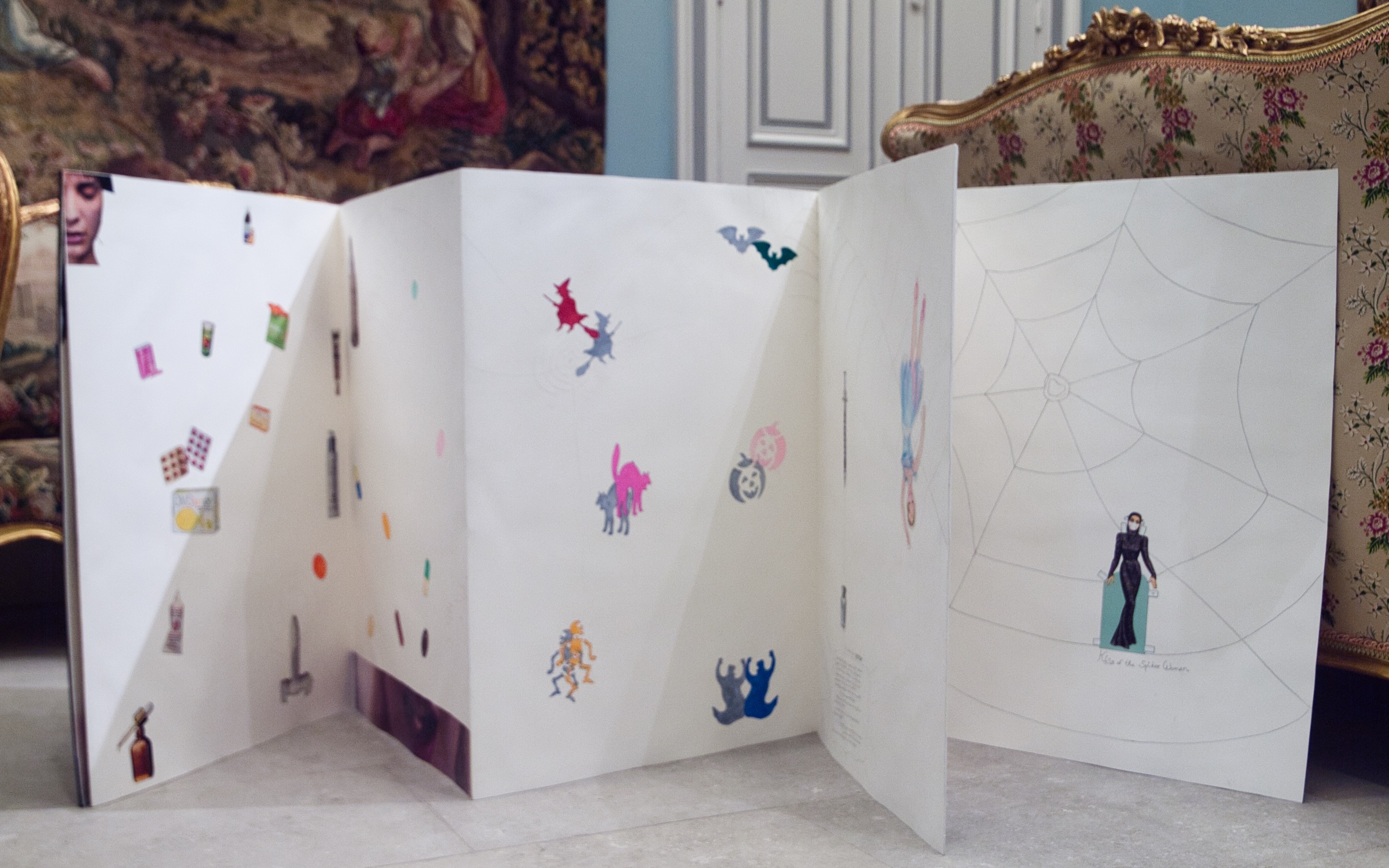 L'image montre un panneau pliable, probablement une sorte d'affichage ou de présentation artistique. Ce dernier est décoré de diverses illustrations et autocollants. On peut y voir des personnages colorés, des motifs fantaisistes, ainsi que des éléments comme des épées et des créatures. À droite, il y a une illustration d'une silhouette féminine vêtue d'une robe noire, entourée d'une toile d'araignée dessinée. L'ensemble est monté sur fond blanc, et le tout semble créer une atmosphère imaginative et ludique.