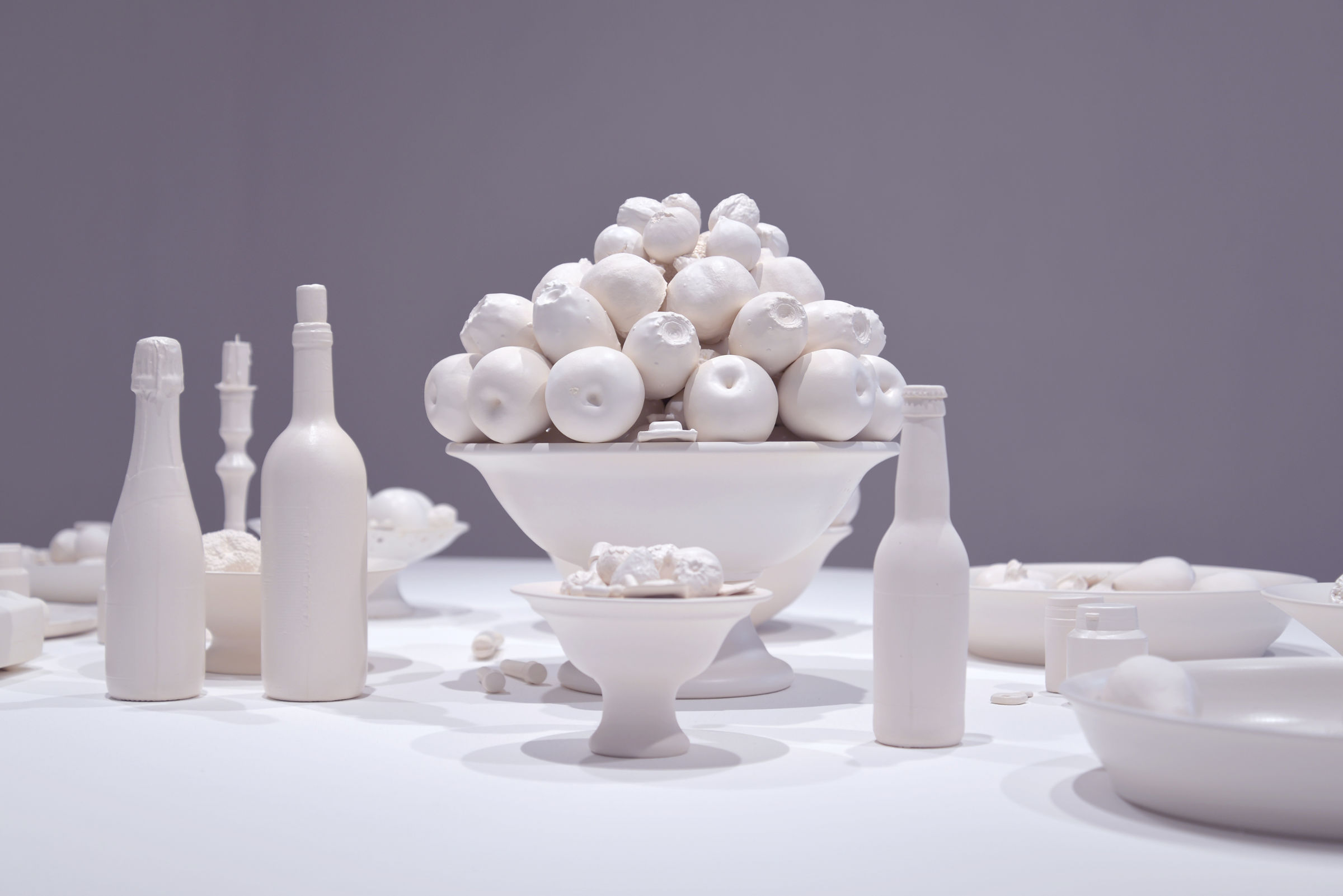 L'image présente une composition artistique minimaliste mettant en scène divers objets en céramique blanche. Au centre, on observe un grand bol surmonté d'une pile de petites pommes blanches. Autour, plusieurs autres récipients tels que des bols et des bouteilles, également en blanc, créent une ambiance harmonieuse et épurée. L'ensemble est disposé sur une surface blanche, ce qui accentue le contraste et donne une impression de simplicité et de sérénité. Le fond gris clair contribue à mettre en valeur les formes et les textures des objets.
