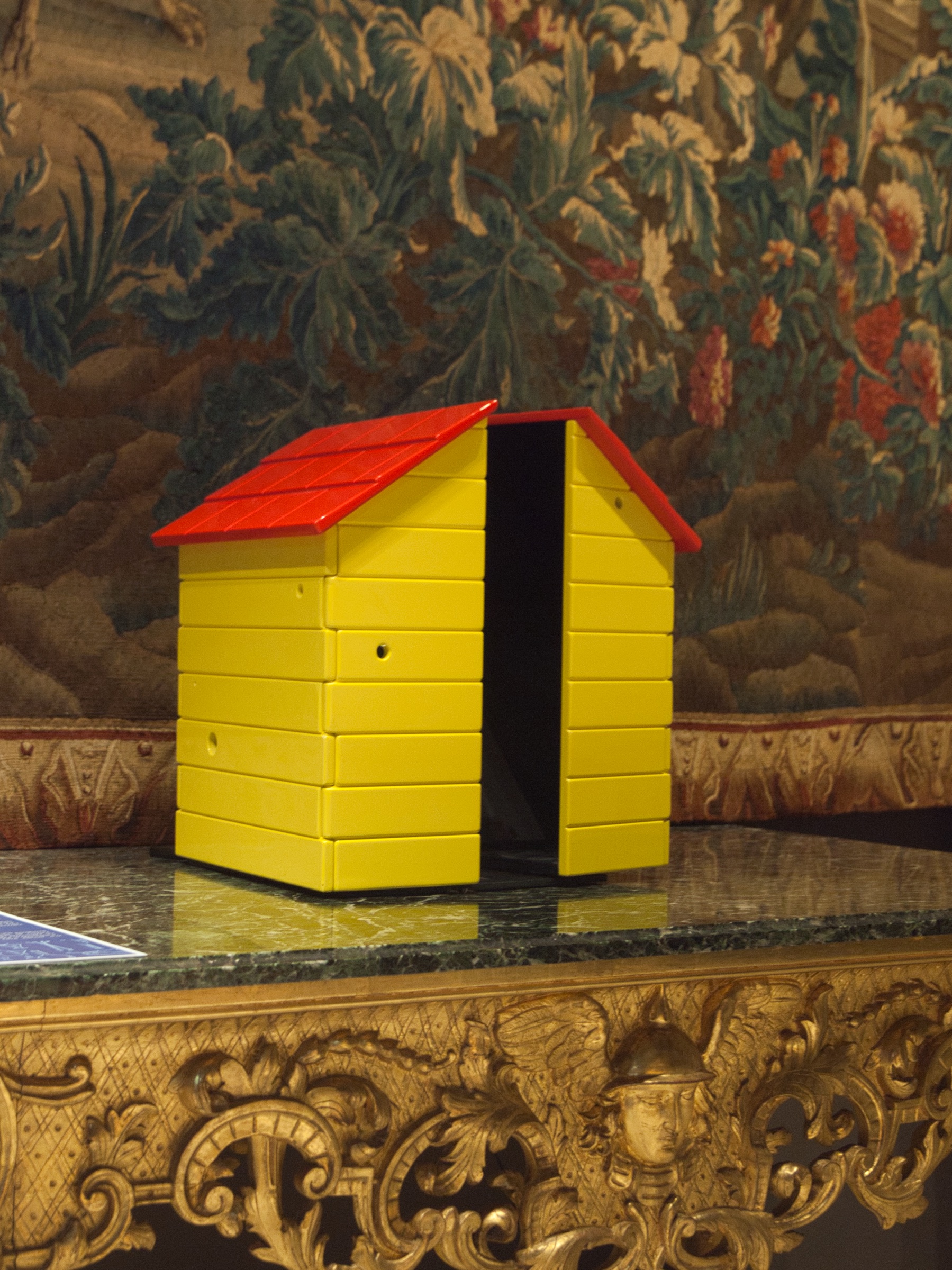 L'image montre une petite maisonnette en bois peinte en jaune avec un toit rouge. Elle est posée sur une surface de marbre, ornée de motifs dorés. En arrière-plan, on aperçoit une tapisserie richement décorée, remplie de motifs floraux et de couleurs variées. La maisonnette semble simple mais attrayante, ajoutant une touche de couleur à l'environnement.