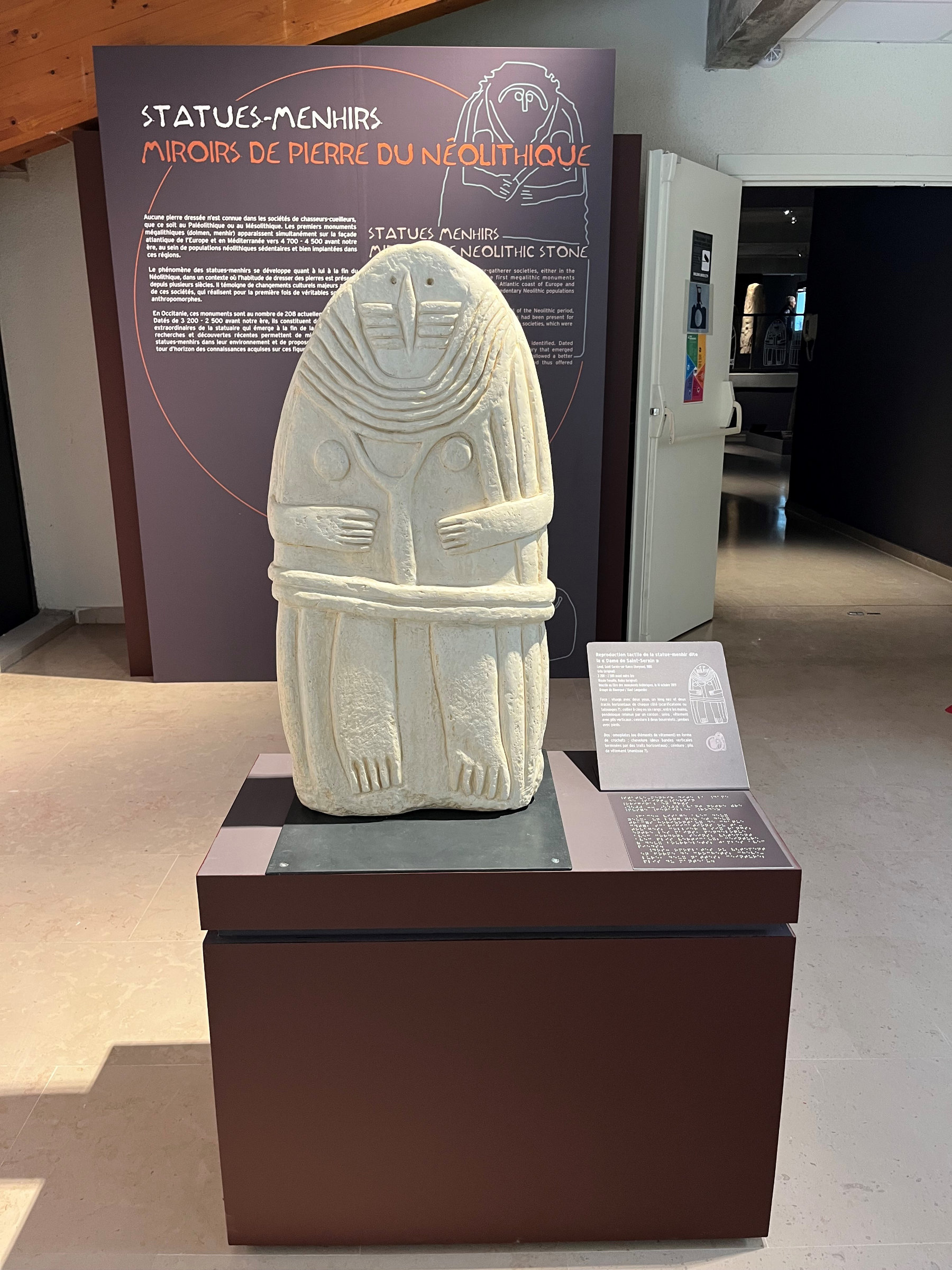 L'image montre une statue en pierre représentant un menhir du Néolithique. La sculpture est placée sur un socle dans un musée, entourée d'une présentation informative. La statue a une forme humanoïde, avec des détails gravés qui lui confèrent une apparence stylisée. Le fond est sombre, ce qui met en valeur la statue claire. La signalétique explique son importance historique, probablement liée aux pratiques et croyances de cette époque.