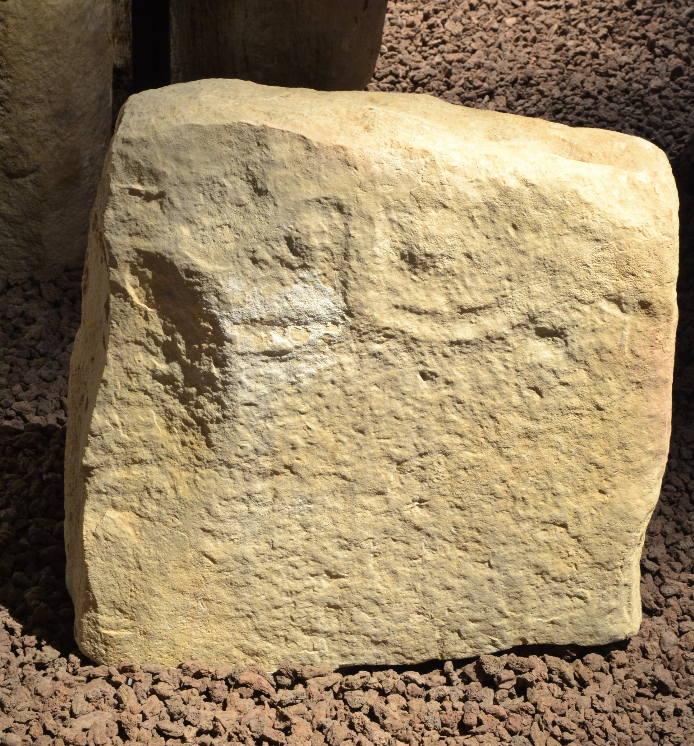L'image montre un gros bloc de pierre de couleur beige ou jaunâtre, possédant une surface rugueuse et irrégulière. Ce bloc est posé sur un sol composé de petits graviers. On peut apercevoir des marques ou des gravures sur la pierre, indiquant un certain niveau de travail ou de sculpture. L'éclairage met en valeur les textures de la surface, renforçant son aspect ancien et brut.