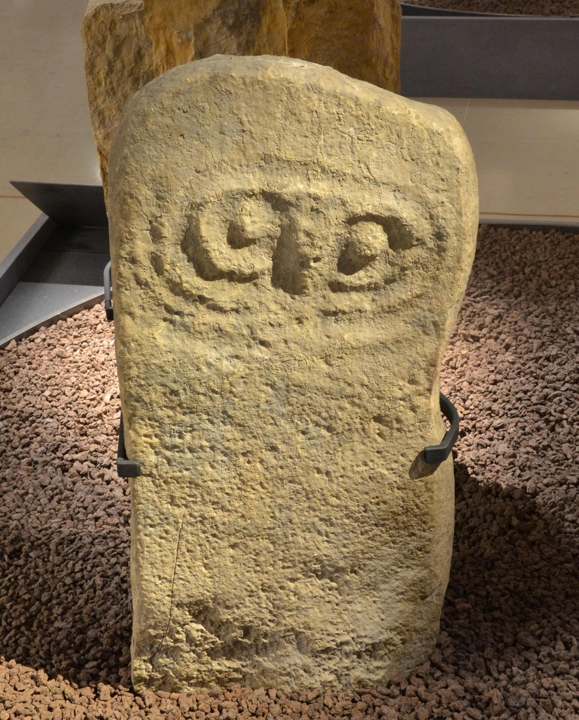 L'image montre un fragment de pierre taillée, probablement une stèle ou une pierre gravée. La pierre a une forme irrégulière et présente des motifs ou des symboles sculptés sur sa surface. Elle repose sur un socle ou un support, entourée de petits graviers. L'aspect général suggère qu'il s'agit d'un objet archéologique, pouvant avoir une signification historique ou culturelle.