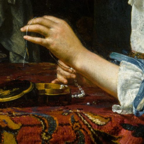 L'image montre une main délicate tenant un fil, probablement en train de travailler sur un collier de perles. Le poignet est orné d'un ruban bleu et la main semble concentrée sur l'activité. Une boîte ou un objet rond en bois est visible sur la table, ainsi qu'une étoffe ornée de motifs richement colorés, créant une atmosphère élégante.