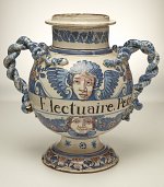 L'image montre un vase décoratif en céramique, caractérisé par ses deux poignées en forme de cordes. Le vase est orné de motifs colorés, notamment des visages d'angelots, des motifs floraux et des couleurs vives, principalement en bleu. Au centre, on peut lire le mot "Electuaire" suivi de "Peto", ce qui suggère un usage en lien avec la pharmacologie ou les remèdes à l'époque. Le style de l'objet évoque une période historique, probablement liée à la céramique européenne, ornée de détails minutieux.