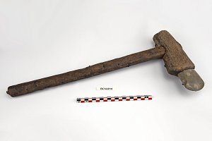 L'image montre un outil qui ressemble à un maillet. Il est constitué d'un long manche en bois, avec une tête en pierre fixée à une extrémité. Le bois semble ancien et usé, ce qui indique qu'il a pu être utilisé pendant une longue période. À côté de l'outil, il y a une règle ou un échelon coloré, probablement utilisée pour donner une échelle à la photo ou pour des fins de mesure. L'arrière-plan est neutre, mettant en avant l'outil.