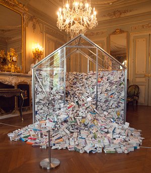 L'image montre une grande structure en forme de serre en verre, remplie de milliers de boîtes de médicaments. Cette installation est située dans une pièce élégante, ornée de miroirs et de luminaires dorés, avec un sol en parquet. Les boîtes de médicaments sont entassées à l'intérieur de la serre, créant un effet visuel impressionnant, contrastant avec le décor luxueux de la pièce.