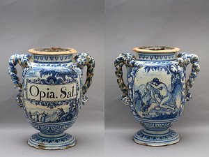 L'image montre un grand vase en céramique décoré de motifs en bleu et blanc. Le vase a deux anses ornées, et on peut distinguer des scènes peintes représentant des figures humaines. Sur une des faces, une inscription est visible, mentionnant "Opia. Sal." Les détails et les motifs ajoutent une touche artistique, mettant en valeur le style classique de la poterie. Le vase semble ancien et témoigne d'un savoir-faire artisanal.