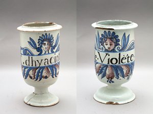 L'image montre deux poteries décoratives, probablement des vases ou des contenants. Chaque pot est orné de motifs floraux colorés, avec des visages stylisés intégrés dans la décoration. Le premier pot porte le nom "C. d'hyacinthe" et le second "e. Violette", suggérant qu'ils sont associés à des fleurs spécifiques. Les couleurs utilisées incluent des nuances de bleu, vert et des détails en brun, ce qui donne un aspect artisanal et vibrant. Les bords des pots semblent légèrement usés, indiquant qu'ils ont été utilisés ou exposés pendant un certain temps.