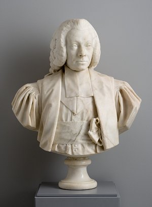 L'image représente un buste sculpté en marbre. Le buste montre un homme avec des cheveux bouclés et portant des vêtements d'époque, notamment une chemise avec de larges manches et un gilet. Il a une expression sérieuse et est représenté de manière réaliste. Le buste est présenté sur un socle cylindrique, et l'arrière-plan est d'une couleur neutre, ce qui met en valeur le marbre et les détails de la sculpture.