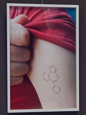 L'image représente un bras humain portant un tatouage. Le bras est vêtu d'un t-shirt rouge, et le tatouage représente une structure chimique composée de plusieurs hexagones, qui peuvent évoquer une molécule. Cette illustration évoque probablement un lien avec la science ou la chimie, et le contraste entre le t-shirt coloré et la ligne délicate du tatouage attire l'attention. On voit une main qui tient le bras, ce qui donne un aspect personnel à l'image.