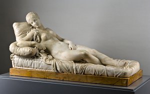 L'image représente une sculpture en marbre d'une femme allongée, dépeignant une figure féminine nue. Elle repose sur un coussin avec une pose détendue, les mains posées sur le corps et un regard apaisé. La sculpture est finement détaillée, mettant en avant la texture des drapés autour d'elle ainsi que les traits délicats de son visage. Le tout est présenté sur une base en marbre qui accentue la beauté de l'œuvre.