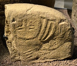 Cette image montre une pierre avec des inscriptions ou des gravures anciennes. La surface de la pierre semble rugueuse et il y a des motifs sculptés qui pourraient représenter des symboles ou des figures. La pierre est exposée dans un environnement muséal, entourée de petits gravillons. L'éclairage met en valeur les détails des gravures, suggérant qu'il s'agit d'un objet d'importance historique ouarchéologique.