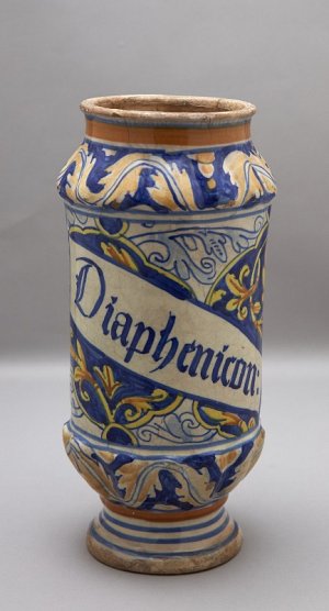 Cette image montre un vase en céramique décoré de motifs colorés. Le fond présente des tons de bleu et d'orange, avec des motifs floraux élaborés. Au centre, une inscription est visible, écrite en lettres dorées sur un fond clair, portant le mot "Diaphenicon". Le vase a une forme élancée et une base plus large, ce qui lui confère une belle stabilité. Les détails de la décoration semblent raffinés et montrent un artisanat soigné.