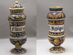 L'image montre deux vases ornés de motifs colorés. Les vases sont de forme élancée, avec une base plus large et un couvercle sur l'un d'eux. Ils présentent des décorations complexes en bleu, orange et jaune, ainsi que des inscriptions en lettres blanches sur un fond coloré. Le premier vase mentionne "Filoni" et le second "D. Agrippa". Les motifs décoratifs incluent des éléments floraux et géométriques, donnant une impression d'artisanat traditionnel.