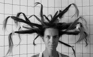 L'image montre une femme posant devant un fond à carreaux. Ses cheveux sont stylisés de manière extravagante, formant des mèches qui s'étendent dans différentes directions, créant une apparence presque sculpturale. Les cheveux sont en partie fixés avec des bandes adhésives, ce qui accentue le côté artistique et original de la coiffure. L'image est en noir et blanc, ce qui donne une atmosphère graphique et dramatique à la composition. Le regard de la femme est sérieux et captivant.