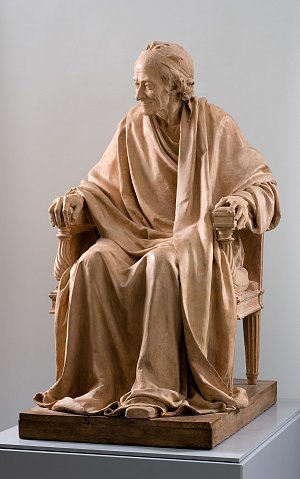 Cette image montre une sculpture en terre cuite représentant un homme assis, vêtu d'une robe ample. Il est placé sur une chaise avec un dossier, ses mains reposant sur les bras de la chaise. Le visage de la sculpture est expressif, affichant une certaine sagesse et sérénité. Les détails de la robe et de la chevelure sont soigneusement réalisés, donnant une impression de réalisme. L'ensemble de la sculpture évoque une attitude réfléchie et contemplative.