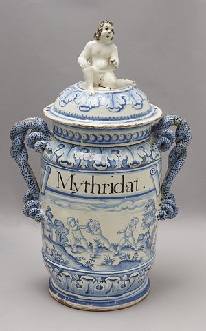 Cette image représente un grand pot ou vase en céramique, décoré de motifs bleu et blanc. Le couvercle est surmonté d'une figurine représentant un personnage nu. Sur le corps du vase, le mot "Mythridat" est inscrit, et se trouvent également des scènes peintes illustrant des motifs animaux et des paysages. Les anses latérales du vase sont ornées de motifs en relief, ajoutant à son caractère décoratif.