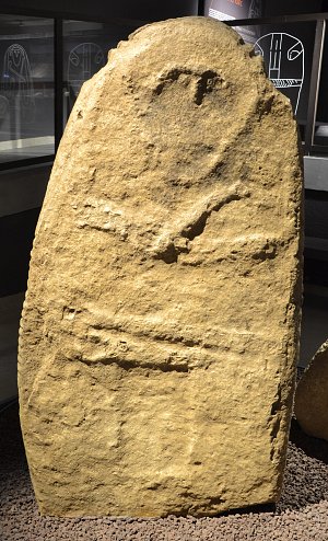 L'image montre une grande pierre taillée, probablement une stèle ou un monument en pierre. La surface de la pierre présente des gravures et des inscriptions qui semblent anciennes. La roche est de forme rectangulaire, avec un sommet arrondi. Le fond est relativement brut, ce qui pourrait indiquer qu'elle provient d'une carrière. L'environnement semble être un espace muséal, avec d'autres artefacts visibles en arrière-plan.