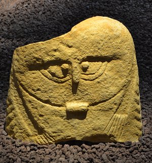 L'image montre une pierre sculptée de couleur jaune, qui semble représenter un visage stylisé. Les traits du visage, tels que les yeux et la bouche, sont dessinés de manière simplifiée et géométrique. La texture de la pierre est rugueuse, et elle se tient sur un fond de granulés foncés, ce qui souligne sa couleur vive. L'œuvre pourrait évoquer un style artistique ancien ou une culture spécifique.