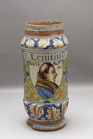 L'image montre un récipient en céramique ornée de motifs colorés. Le vase, de forme cylindrique, présente des décorations complexes, notamment des fleurs et des motifs géométriques. Sur un côté, il y a un portrait en profil d'un homme, probablement un personnage historique ou religieux, accompagné d'inscriptions. Les couleurs utilisées sont vives et varient entre le bleu, le jaune et le vert, ajoutant à l'esthétique générale de l'objet.