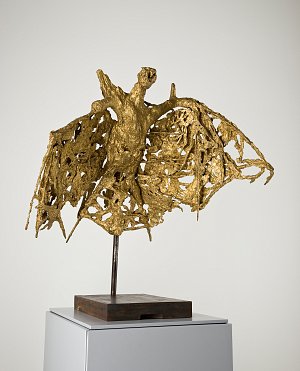 L'image représente une sculpture qui semble évoquer une figure humanoïde avec des ailes, réalisée dans un matériau doré. La sculpture présente une texture à la fois complexe et fluide, avec des formes organiques qui semblent se déployer et se mêler, créant un effet dynamique. Elle est placée sur un socle sobre, ce qui met en valeur son design artistique et sa hauteur. L'ensemble dégage une impression de mouvement et une esthétique moderne.