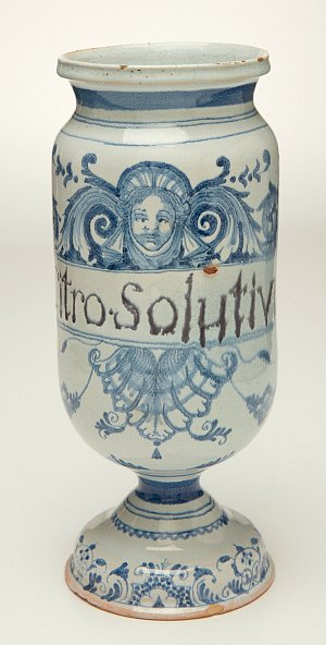 L'image montre un vase en céramique, décoré de motifs en bleu sur fond blanc. Il a une forme élancée avec un pied et un corps arrondi. Des figures ornementales, ainsi que des visages stylisés, sont discernables sur la surface. On peut également voir un texte, "Nitro Solutivo", inscrit sur le vase, suggérant qu'il pourrait avoir eu une fonction particulière ou symbolique. Ce type d'objet évoque une période historique, souvent associée à des artisans spécialisés dans la poterie.