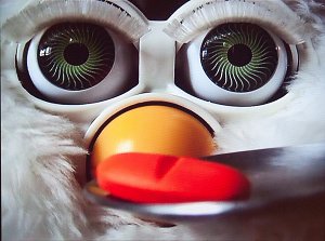 L'image montre un gros plan d'un jouet, probablement un Furby, avec de grands yeux ronds aux iris verts tourbillonnants. Le jouet a un bec orange et se trouve devant une cuillère contenant un comprimé rouge. L'expression du jouet, avec son regard fixe, crée une atmosphère intriguante.