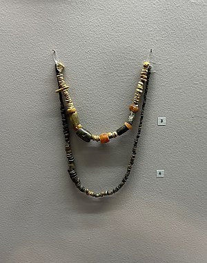 L'image présente un collier ou une chaîne ornée de plusieurs perles disposées en différentes formes et couleurs. Les perles semblent être fabriquées à partir de matériaux variés, avec des textures et des motifs distincts. Le collier est suspendu à un mur gris, et on peut voir des étiquettes numérotées à côté de lui, probablement pour identifier ou décrire l'objet dans un contexte d'exposition. Les couleurs dominantes semblent être des nuances de noir, doré et brun.