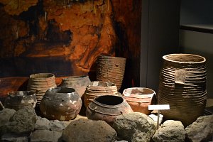 L'image montre une collection de poteries anciennes disposées sur une surface, entourées de pierres. Les pots ont différentes formes et textures, certains sont décorés avec des motifs. L'arrière-plan présente une teinte sombre qui évoque une ambiance archéologique, suggérant que ces objets sont exposés dans un musée ou un site historique. Les poteries illustrent la créativité et l'artisanat des civilisations passées.