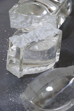 L'image montre plusieurs blocs transparents, en forme de barres, qui ressemblent à du verre ou à de la glace. À l'intérieur de ces blocs, on peut voir une surface lisse avec un liquide clair. La lumière se reflète sur leur surface, créant un effet brillant. Il y a également des morceaux de glace brisée qui semblent être éparpillés autour, ajoutant une texture intéressante à la composition. L'arrière-plan est sombre, ce qui met en valeur la clarté et la transparence des éléments.