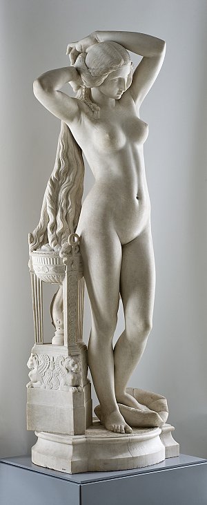 L'image montre une statue en marbre représentant une femme nue. Elle est dépeinte debout, avec un bras au-dessus de sa tête et l'autre le long de son corps. La sculpture semble capturer un moment de grâce et d'élégance, avec des détails raffinés dans la chevelure et les contours du corps. La base de la statue est ornée de motifs décoratifs, ajoutant à son esthétique globale. L'ensemble dégage une impression de beauté classique et de sérénité.